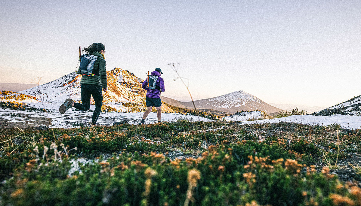 Two hikers with backpacks on a mountain trail with snow-capped peaks in the background.