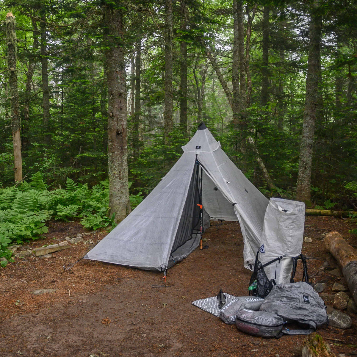 Front exterior view of the Hyperlite Mountain Gear Shelters UltaMid 2 Half Insert inside white tent in a wooded setting. Assorted Hyperlite hiking gear sits beside the outside right of the tent