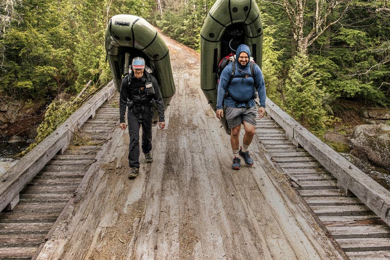 Two people carrying inflatable kayaks on a wooden bridge in a forest setting