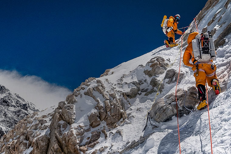 Two climbers in orange jackets ascending a snowy mountain peak with clear blue sky.