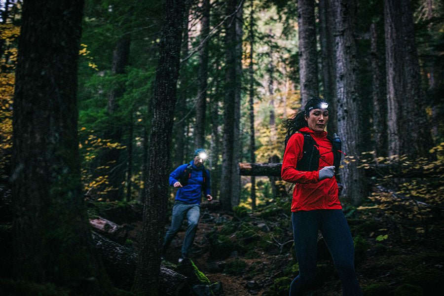 Two people running through a forest at night with headlamps.