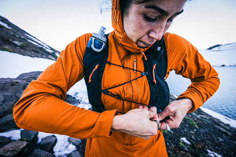 Person in orange jacket with backpack adjusting something on a snowy mountain background