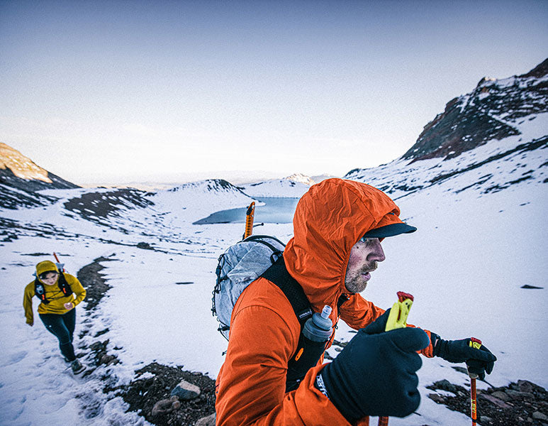 Person in orange jacket hiking in a snowy mountain landscape with another person in the background.