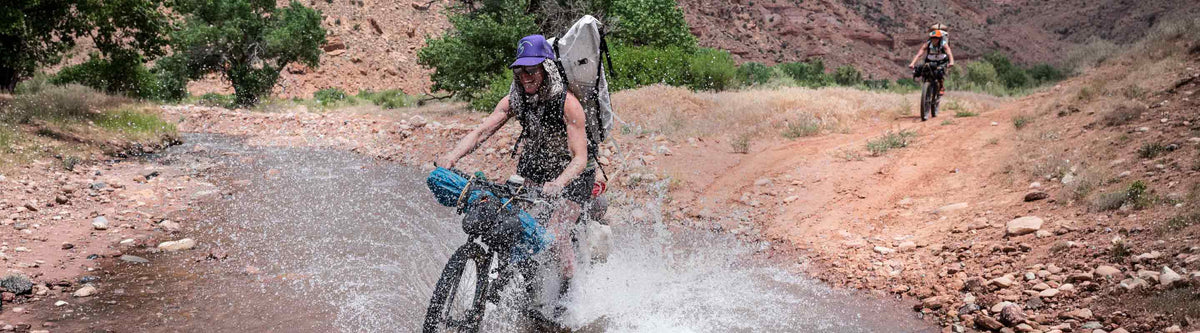Person riding a bike through water on a dirt road with mountains in the background