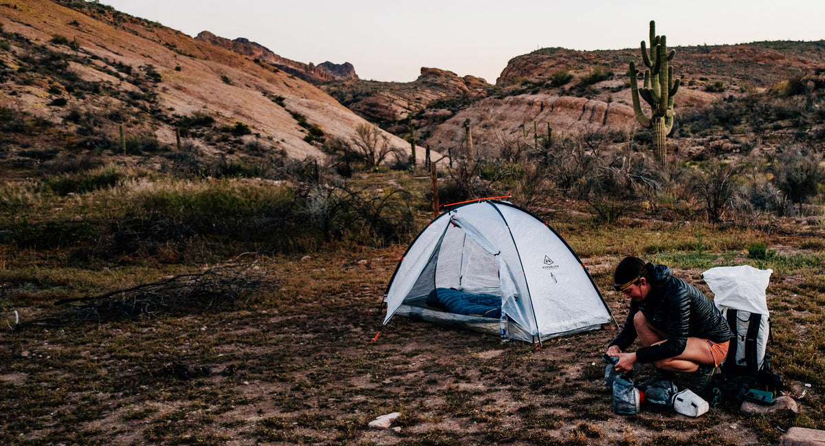 Person bent down near a tent in a desert landscape with cacti and rocky hills.