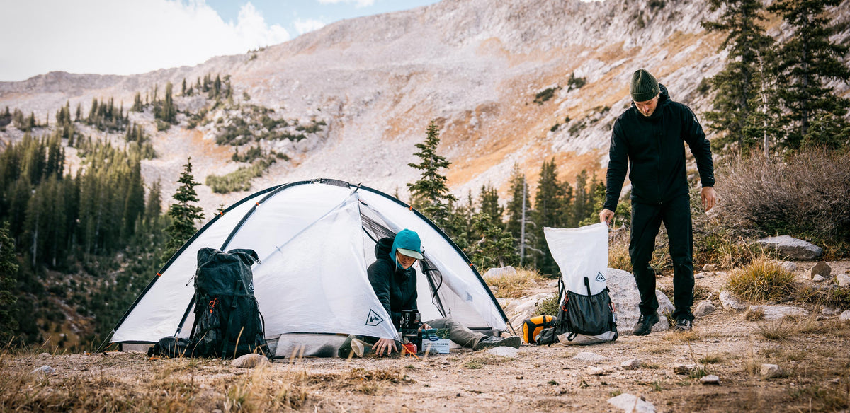 Two people setting up a tent in a mountainous area with backpacks and camping gear.