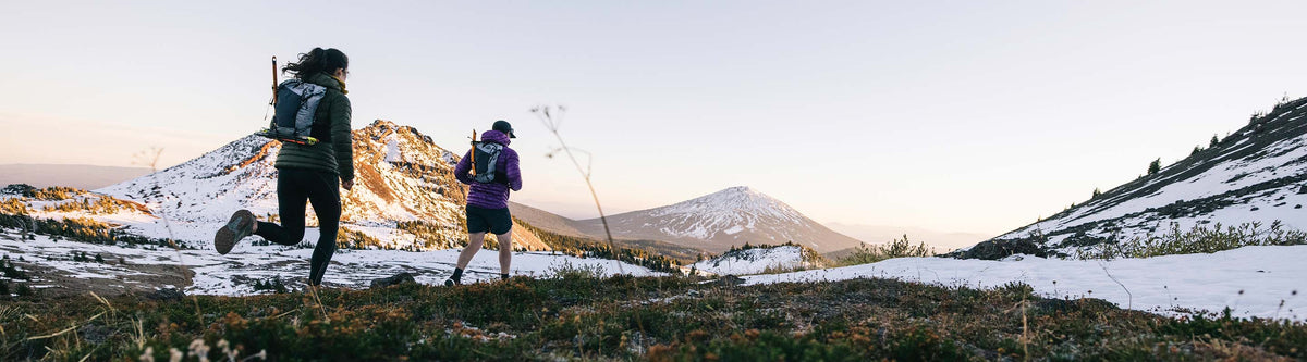 Two hikers with backpacks running on a snow-covered mountain landscape.