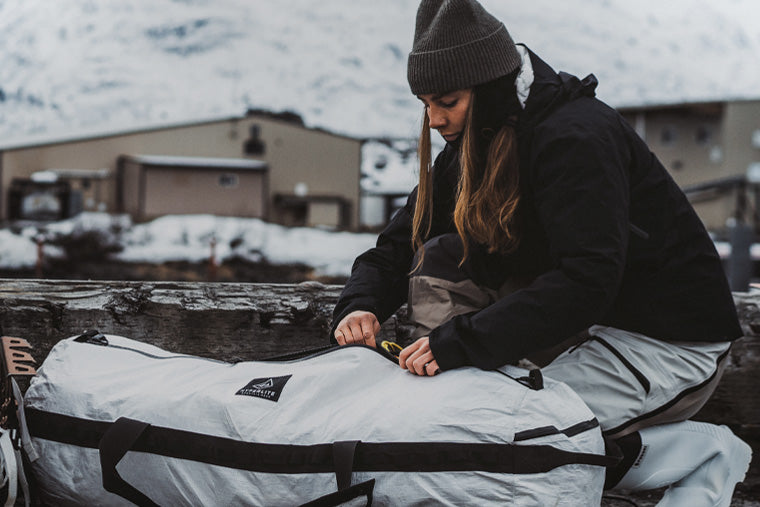 Person in winter clothing preparing a large white bag outdoors with snow-covered mountains in the background.