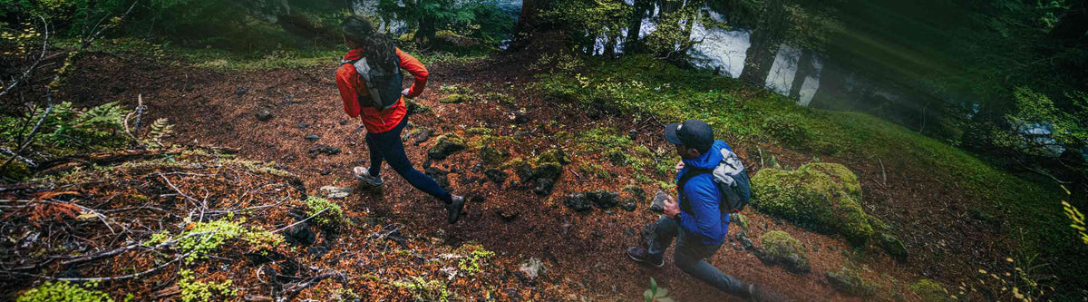 Two hikers with backpacks running through a forest near a body of water.
