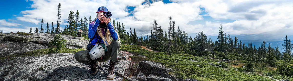 Person crouching down on a rock and p a camera in a mountainous area with trees holding and sky in the background
