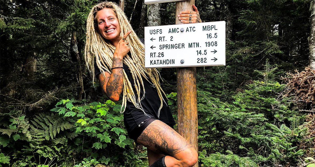 Hiker Liz Kidder smiling and pointing to a trail sign on the Appalachian Trail, marking the long distance to Katahdin and Springer Mountain.