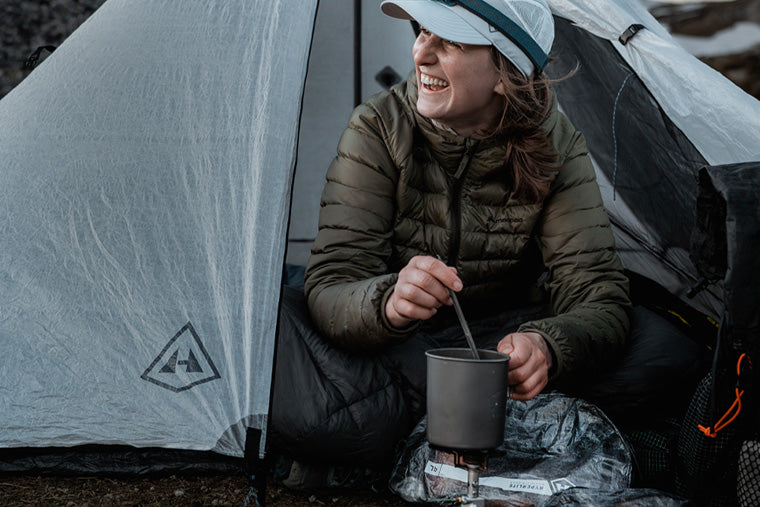 Person sitting outside a tent using a camping stove with a cup.