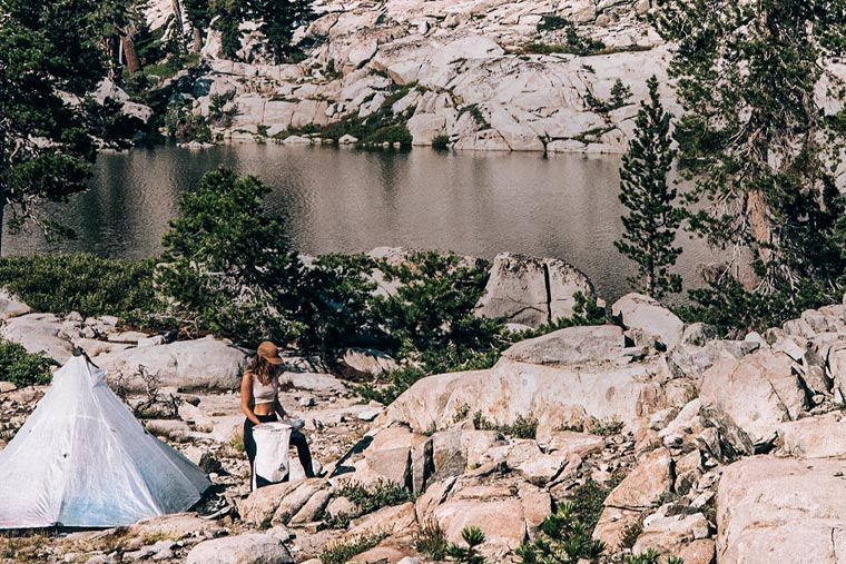 Person standing by a tent near a lake surrounded by rocks and trees