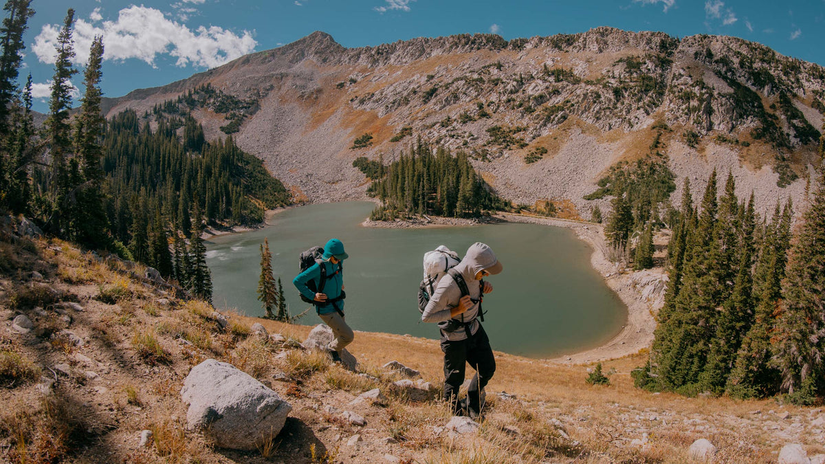 Two hikers with backpacks walking towards a mountain lake on a sunny day.