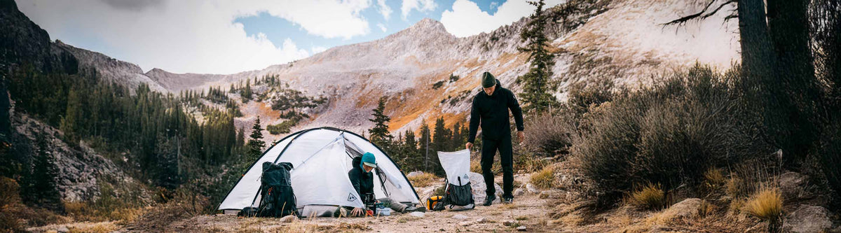 Two people in a camping area with a tent in a mountainous area with trees and rocks.