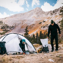 Two people setting up camping gear in a mountainous area with a tent and backpacks.