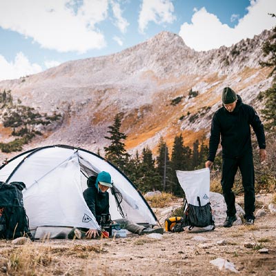 Two people setting up camping gear in a mountainous area with a tent and backpacks.