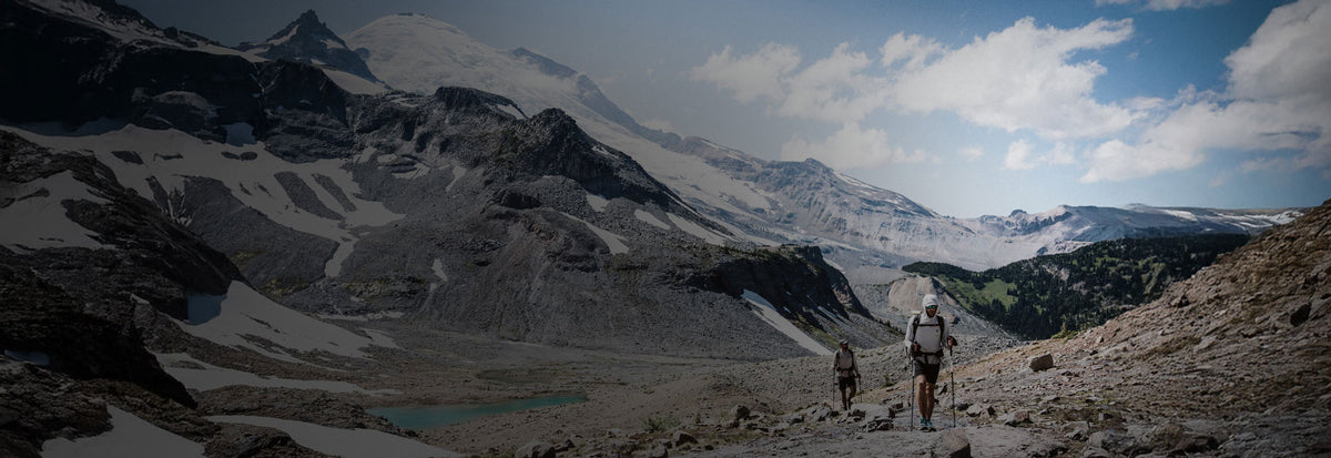 Two hikers on a mountain trail with snow-capped peaks and a clear sky.